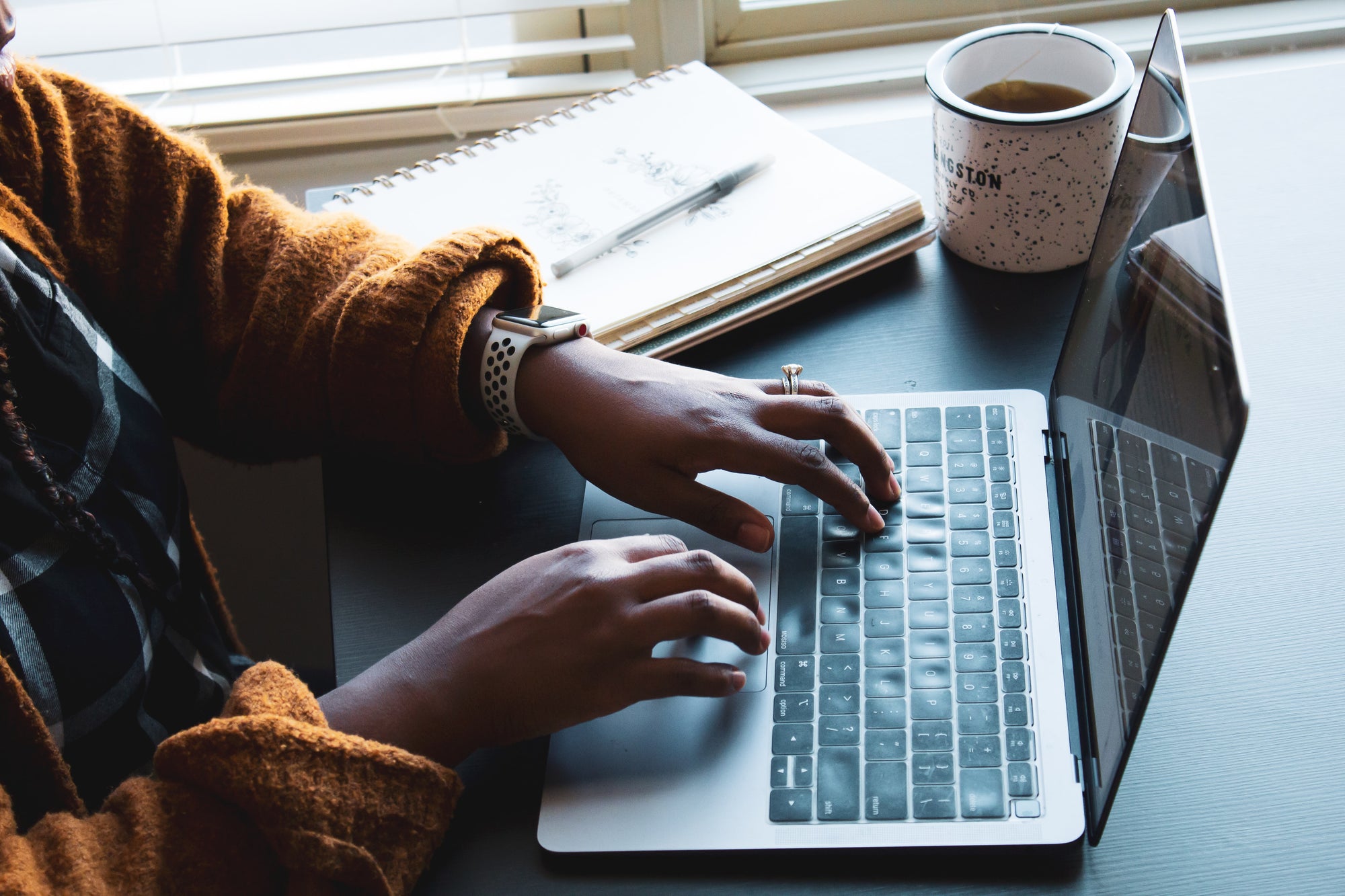 A black woman types on a laptop with coffee and a notebook nearby